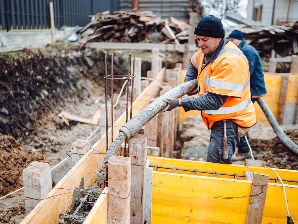A man in a florescent vest and work clothes pumps concrete into a tight space.